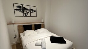 A neatly made bed with white linens and a black pillow, a wooden headboard, and a wall-mounted artwork of a tree.
