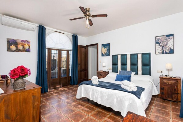Une chambre d’hôtel avec un lit, une commode en bois, un bureau, un ventilateur de plafond et des œuvres d’art accrochées aux murs.