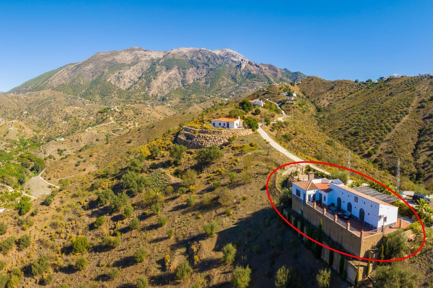 A mountainous landscape with a white building featuring a red roof and a stone wall, surrounded by greenery and a clear sky.