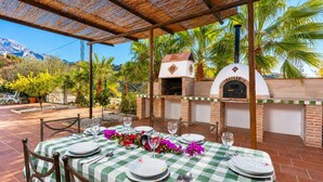 Un espacio de comedor al aire libre con mantel a cuadros, dos hornos de ladrillo y vistas a palmeras y montañas.