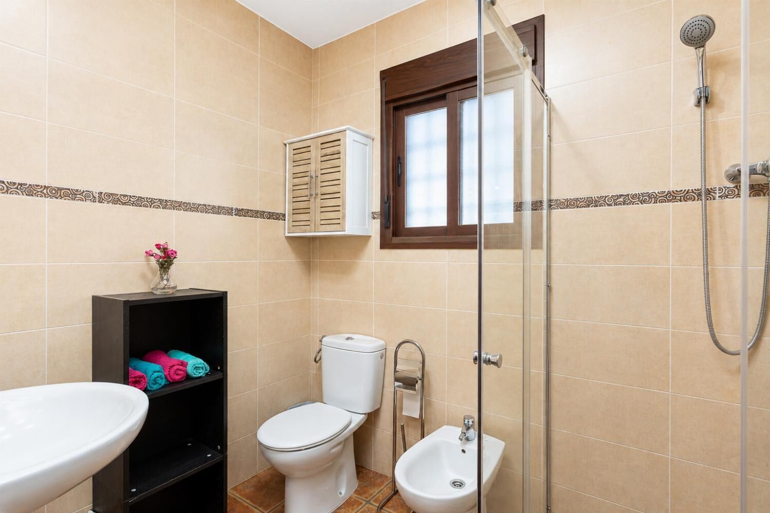 A bathroom with a toilet, bidet, and shower. There is a shelf with towels and a cabinet above the toilet.
