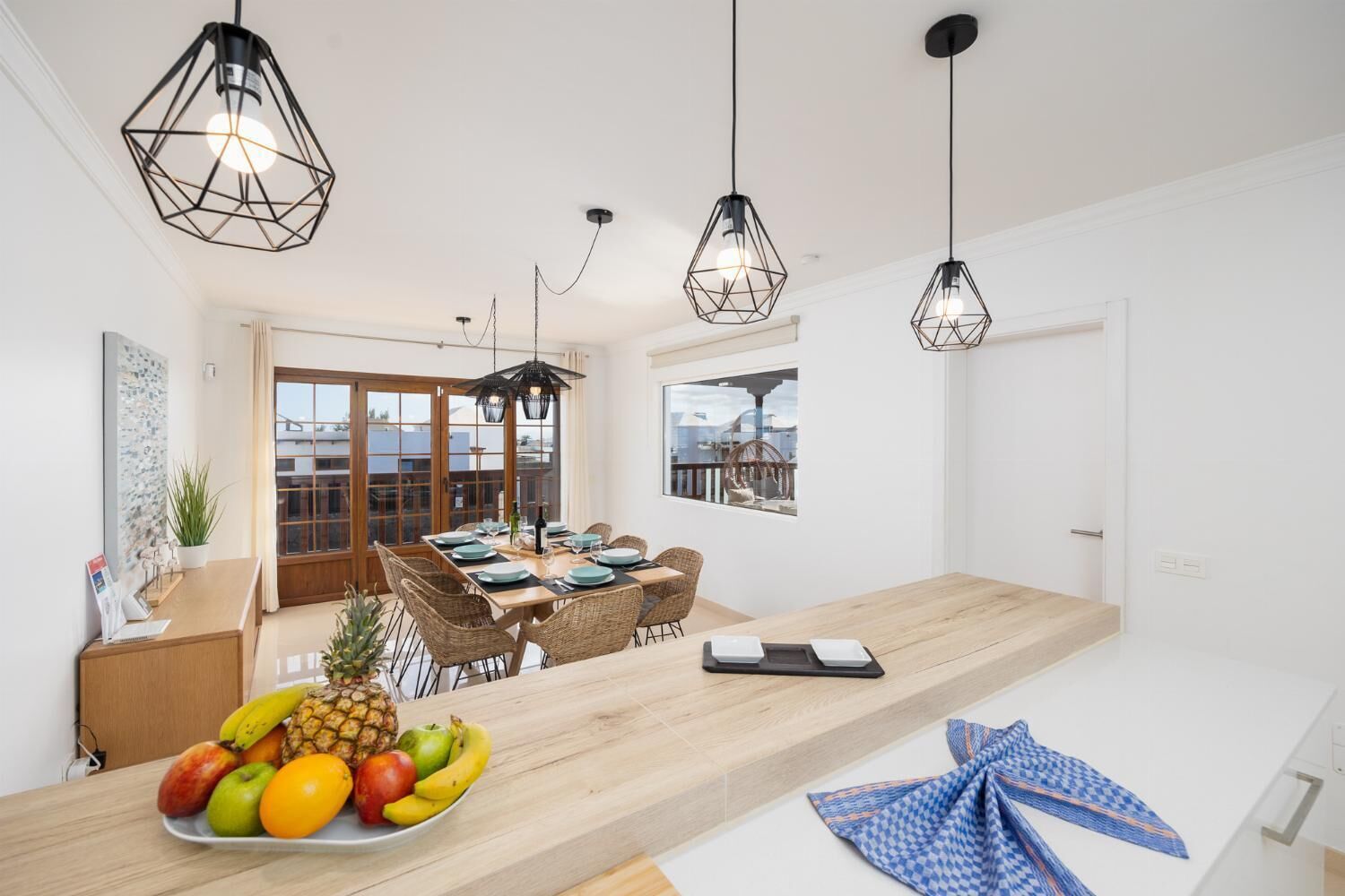 A modern dining area with a wooden table, wicker chairs, and a fruit bowl. There are pendant lights above the table.