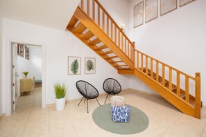 Eine Treppe mit hölzernen Geländern, ein Blumentopf, zwei schwarze Stuhlgestelle aus Draht, ein gemustertes Kissen und gerahmte Blätterkunst an der Wand.
