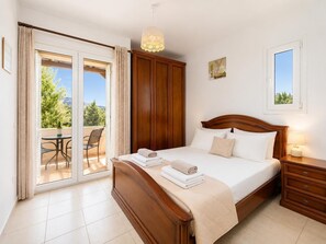 A bedroom with a bed, a nightstand, a wooden wardrobe, a balcony with a table and chairs, and a view of greenery.