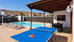 Ett poolområde med ett bordtennisbord, solstolar och en möblerad sittplats under tak.