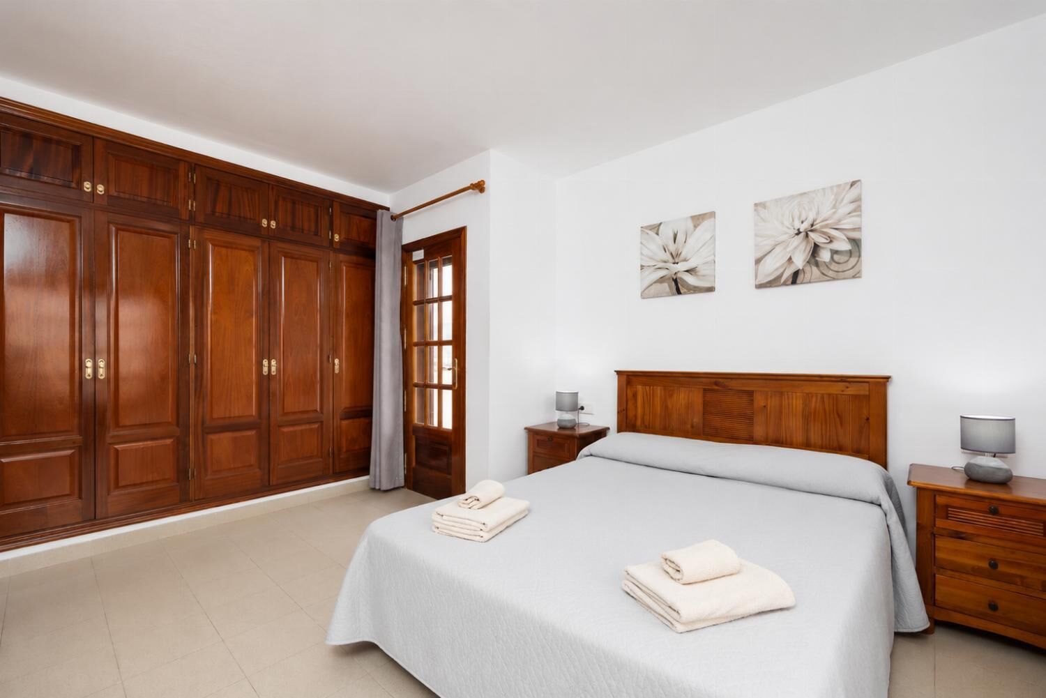Un dormitorio con un armario de madera grande, una cama con cubrecama gris, dos mesitas de noche con lámparas, y dos cuadros de flores colgados en la pared.