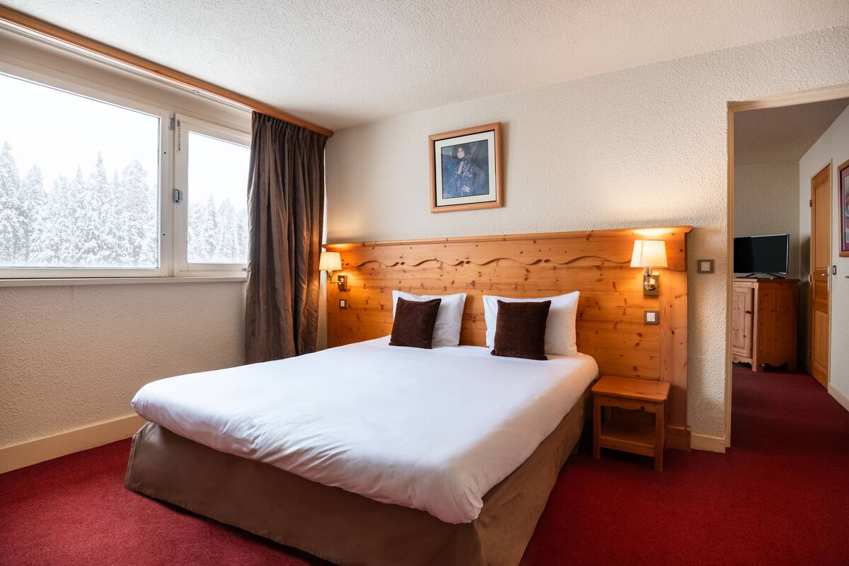 A hotel room with a large bed, wooden headboard, two bedside lamps, a small bedside table, a TV, and a window with a view of snow-covered trees.