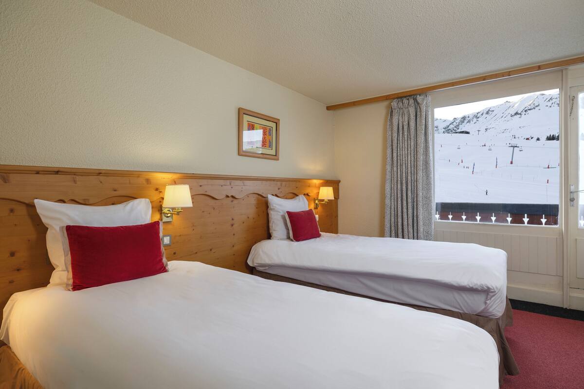 A hotel room with two beds, a wooden headboard, a window with a view of a snowy mountain and ski slope, and a framed picture on the wall.