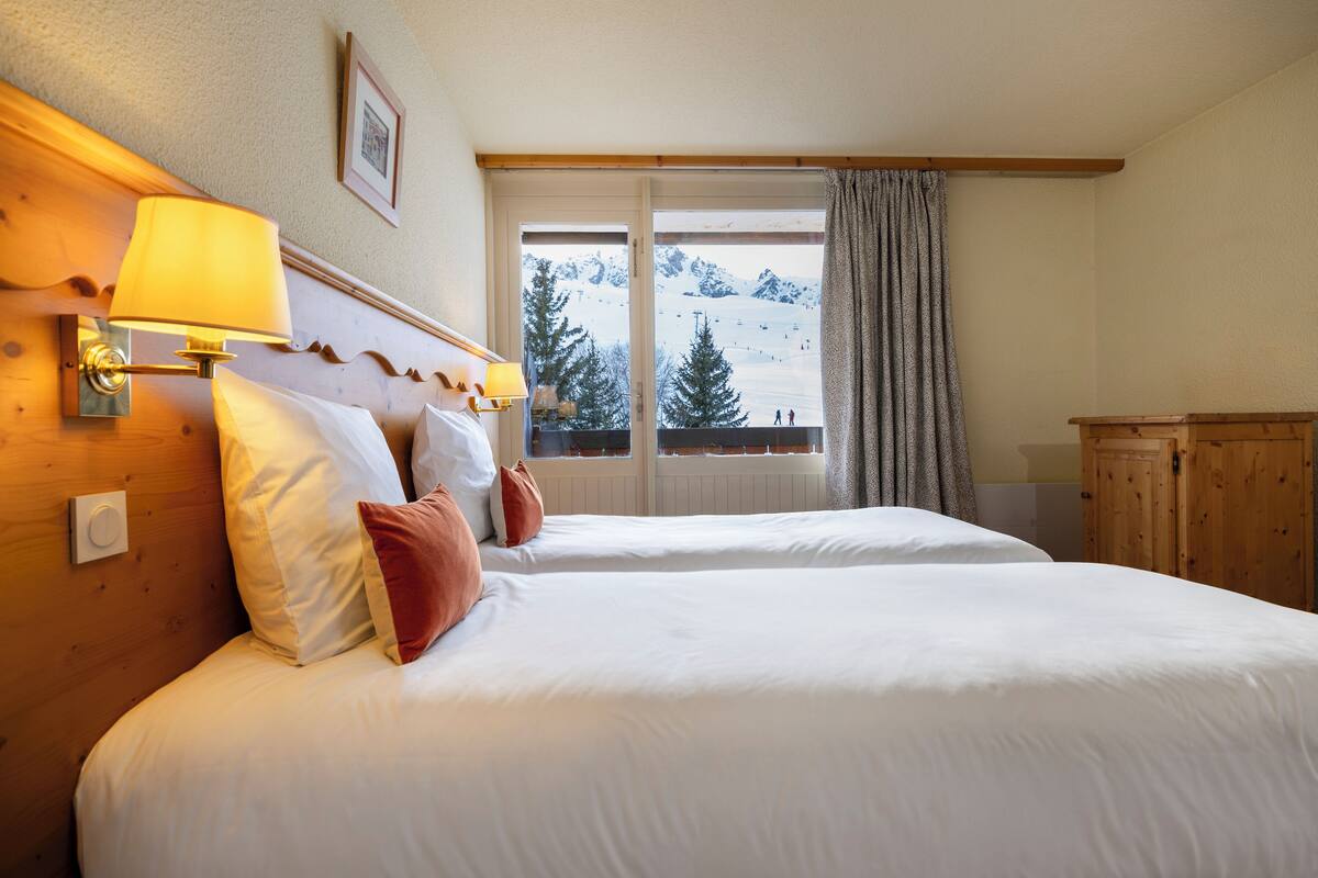A hotel room with two beds, a wooden headboard, a window with a view of a snowy mountain, and a wooden wardrobe.