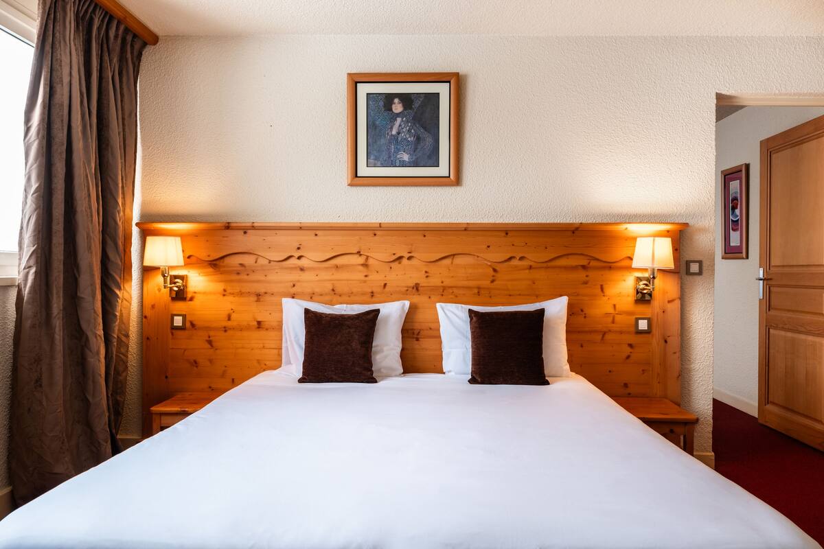 A hotel room with a wooden headboard, two bedside lamps, a framed picture on the wall, and a window with curtains.