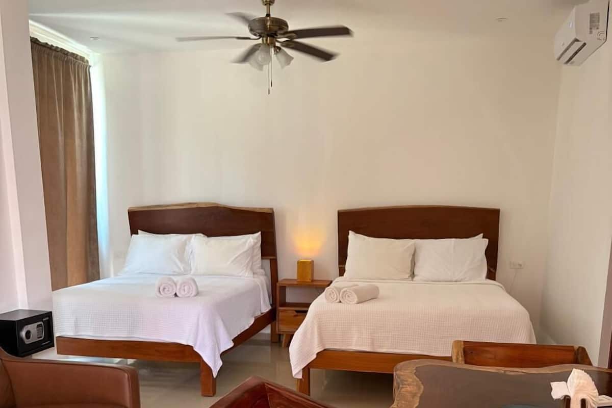 Une chambre d’hôtel avec deux lits, une table basse en bois, un canapé en cuir marron et un ventilateur de plafond.