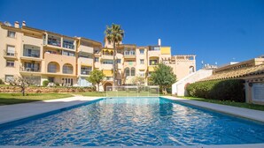 Uma piscina com superfície de água azul cristalina, rodeada por um deck de azulejos brancos. Ao fundo, um edifício de vários andares com varandas e uma palmeira.