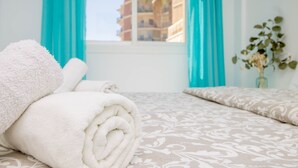 Cama bem arrumada com uma toalha branca enrolada, colcha estampada e um vaso com flores secas num peitoris.