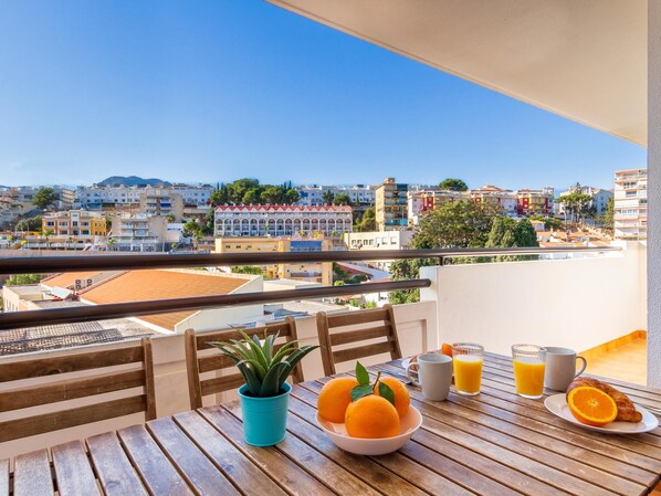 A balcony with a wooden table set for breakfast, including oranges, a croissant, and orange juice.