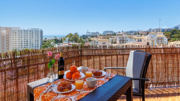 A balcony with a table set for breakfast, including croissants, oranges, and juice, overlooking a cityscape with buildings and a clear sky. - Santa Amalia 51 - Terrace, Swimming Pool, Parking, Sea Views (Torremolinos)