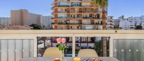 A table with breakfast items, a vase with a flower, and a view of a multi-story building with balconies and palm trees.