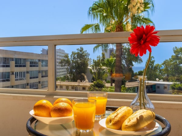Una bandeja de desayuno con naranjas, jugo y pan, un florero con una flor roja, y vistas de edificios y palmeras a través de una ventana.