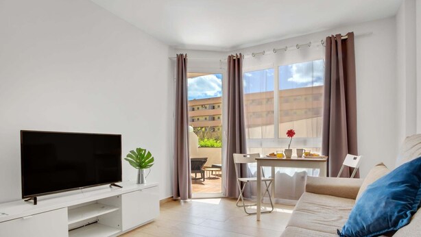 Una sala de estar moderna con televisor de pantalla plana, un sofá, una mesa de comedor con sillas, y un balcón con cortinas.