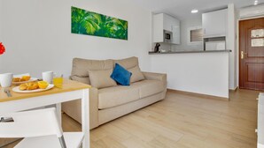 Una sala moderna con un sofá color beige, una mesa de comedor con alimentos para el desayuno, una zona de cocina con armarios blancos y una puerta con un recibo.