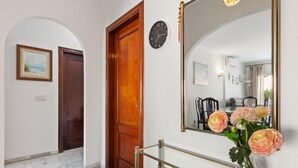 Un couloir avec une porte en bois, une horloge et un miroir qui reflète une salle à manger.