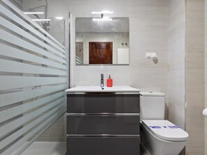 Une salle de bain moderne avec une douche à l’italienne en verre, un lavabo blanc doté d’un distributeur de savon rouge et des toilettes blanches avec une inscription bleue sur le couvercle.