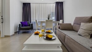 A modern living room with a white coffee table, a sofa, and a dining table set with breakfast items. - Tropico - Beachfront, 1 bedroom, Carihuela (Torremolinos)