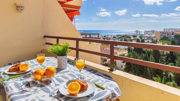 A balcony with a table set for breakfast, including croissants and orange juice, overlooking a coastal cityscape.