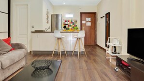 A modern living room with a sofa, a coffee table, and a TV. A kitchen area with a fridge, a sink, and a bar is visible in the background.