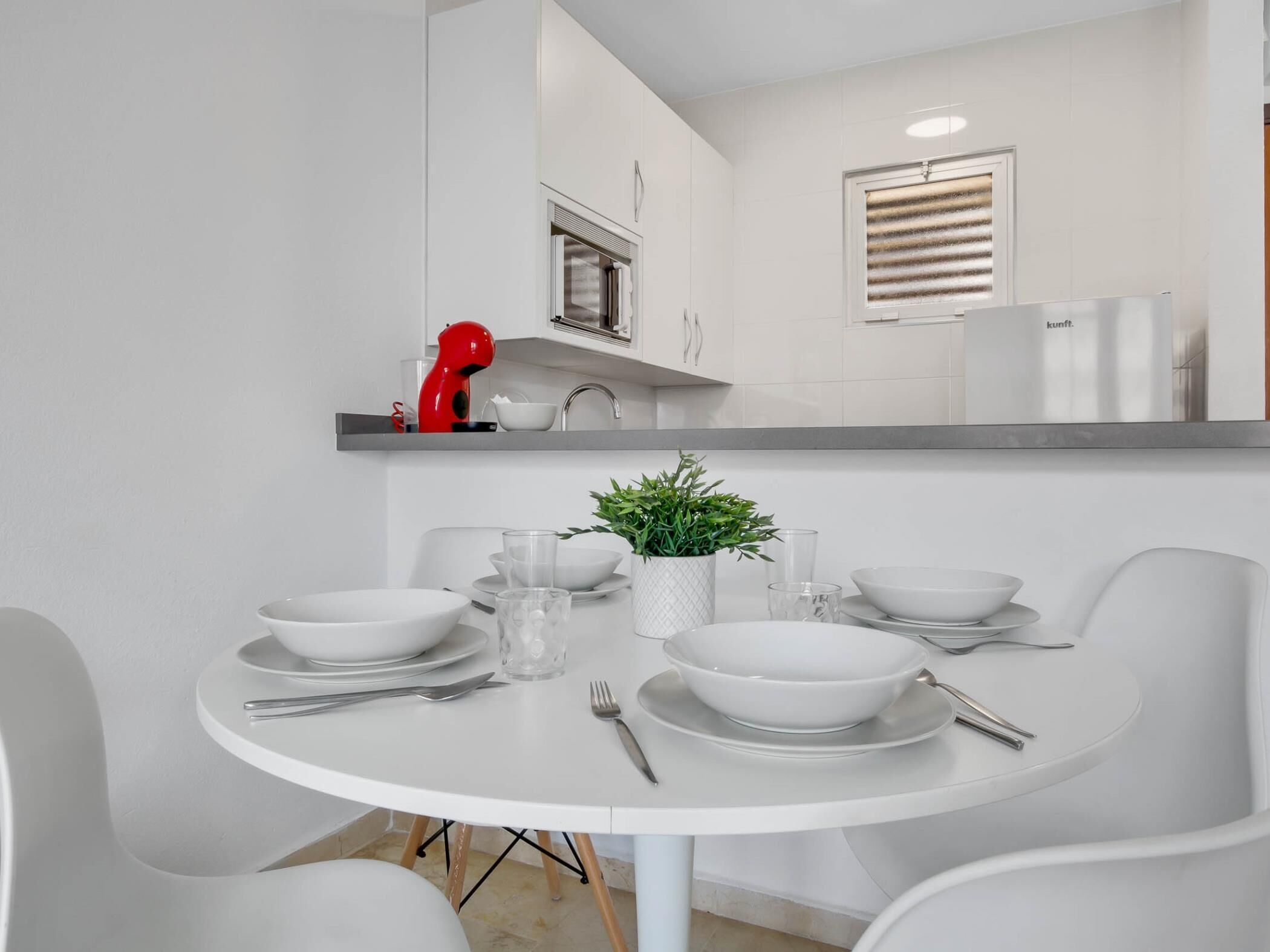 Een moderne keuken met witte kasten, een rode mixer op het aanrecht en een eettafel gedekt met witte borden, kommen en bestek.
