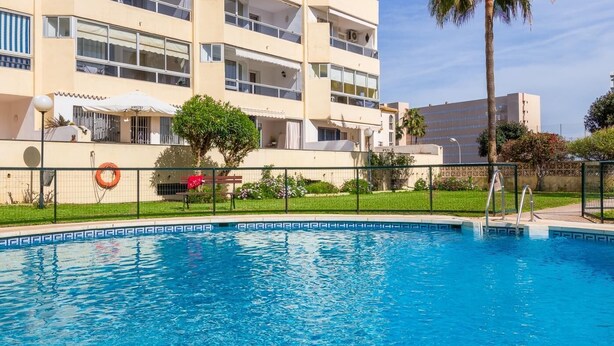 A swimming pool with a clear blue water surface, surrounded by a fence. In the background, there is a multi-story building with balconies and a palm tree. - Vistamarina 405B - Terrace, Swimming Pool, 300m from beach (Torremolinos)