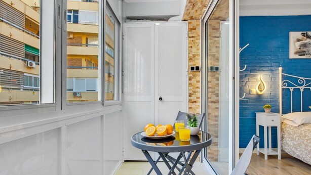 A modern bedroom with a bed, a bedside table, a nightstand, a small table with fruit and drinks, a chair, and a view of a building through a window. - Los Nidos - 1 bedroom, Terrace, 100 m from the beach (Torremolinos)