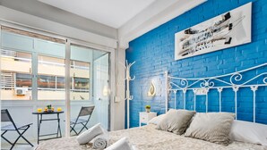 A bedroom with a blue brick wall, a white metal bed frame, and a large window with a view of buildings. - Los Nidos - 1 bedroom, Terrace, 100 m from the beach (Torremolinos)