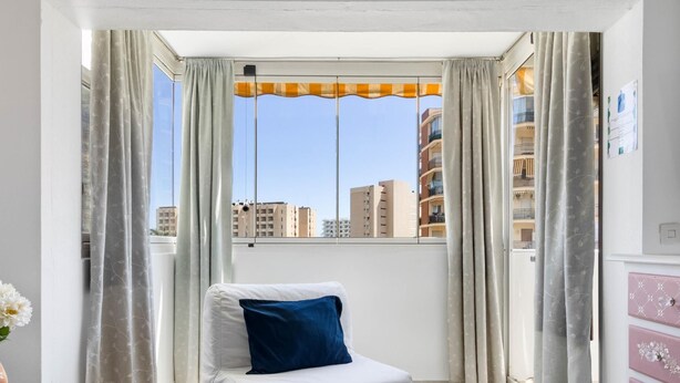 Une pièce avec un canapé blanc, une petite table sur laquelle est posé un vase, et une vue sur des immeubles à travers une fenêtre ornée de rideaux.