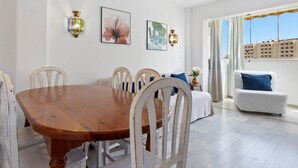Un coin repas avec une table en bois et des chaises blanches, un canapé avec un coussin bleu et deux lampes fixées au mur.