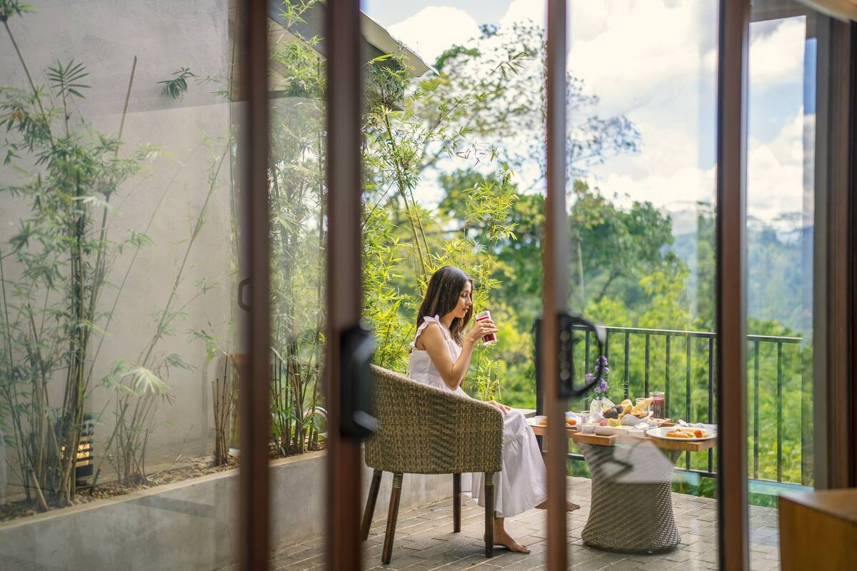 Mulher sentada numa cadeira de vime numa varanda, com uma mesa de comida e bebida, rodeada por plantas de bambu e vista para o verde.