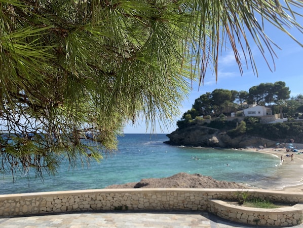 A coastal scene with a stone pathway leading to a beach, a rocky outcrop with buildings, and a clear blue sky.