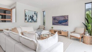 En moderne stue med en hvid sofa, et sofabord og en reol. Der er to billeder på væggen og et stort vindue, der giver naturligt lys.