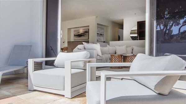 A modern living area with white furniture, a glass door leading to a balcony, and a view of the outdoors.