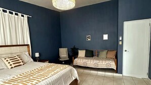A bedroom with a large bed, a smaller sofa, a chair, a bedside table, and a door. - Experience Puerto Rico's Charm in style with modern comforts.  (Luyando)