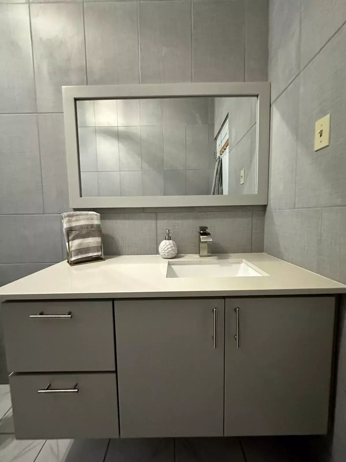 Een badkamer met een grijze betegelde muur, een witte wastafel, een handdoek en een zeepdispenser.