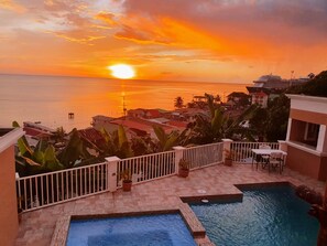 Ein Poolbereich mit Blick auf den Sonnenuntergang über dem Meer, umgeben von Gebäuden und Palmen.