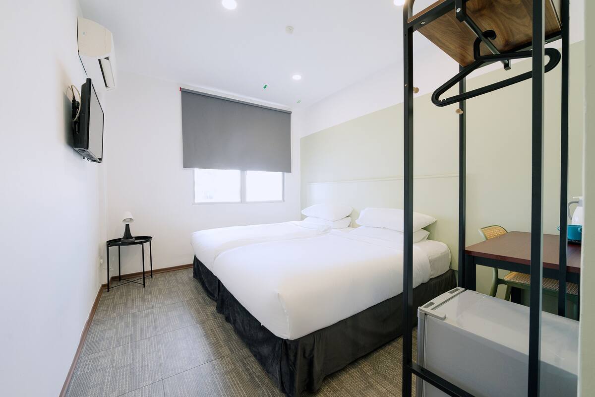 Une chambre d’hôtel avec un lit, une petite table avec une lampe, une fenêtre avec un store et un miroir avec un cadre en bois.
