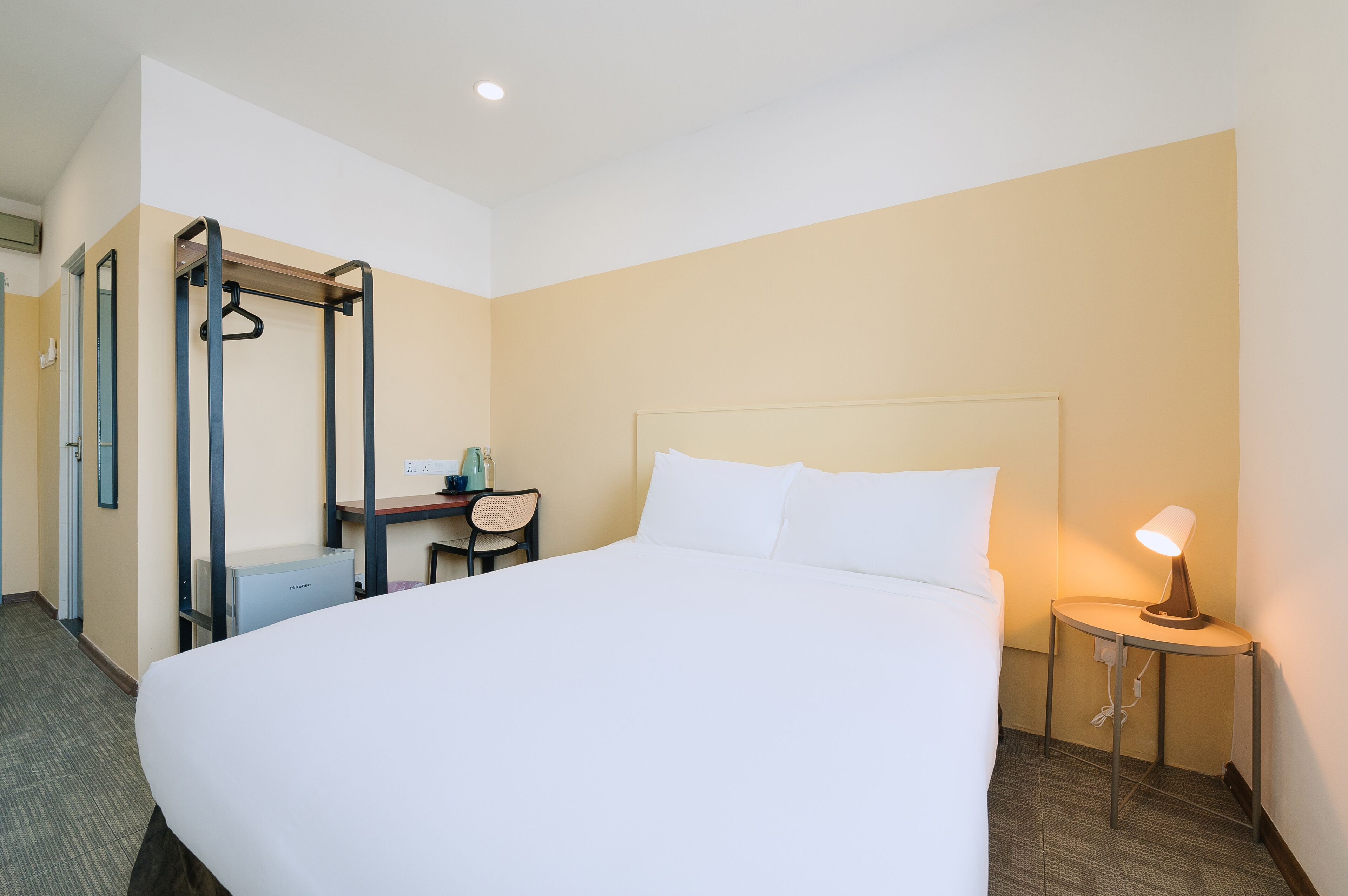 A hotel room with a large bed, a desk with a chair, a bedside table with a lamp, and a wall-mounted radiator.