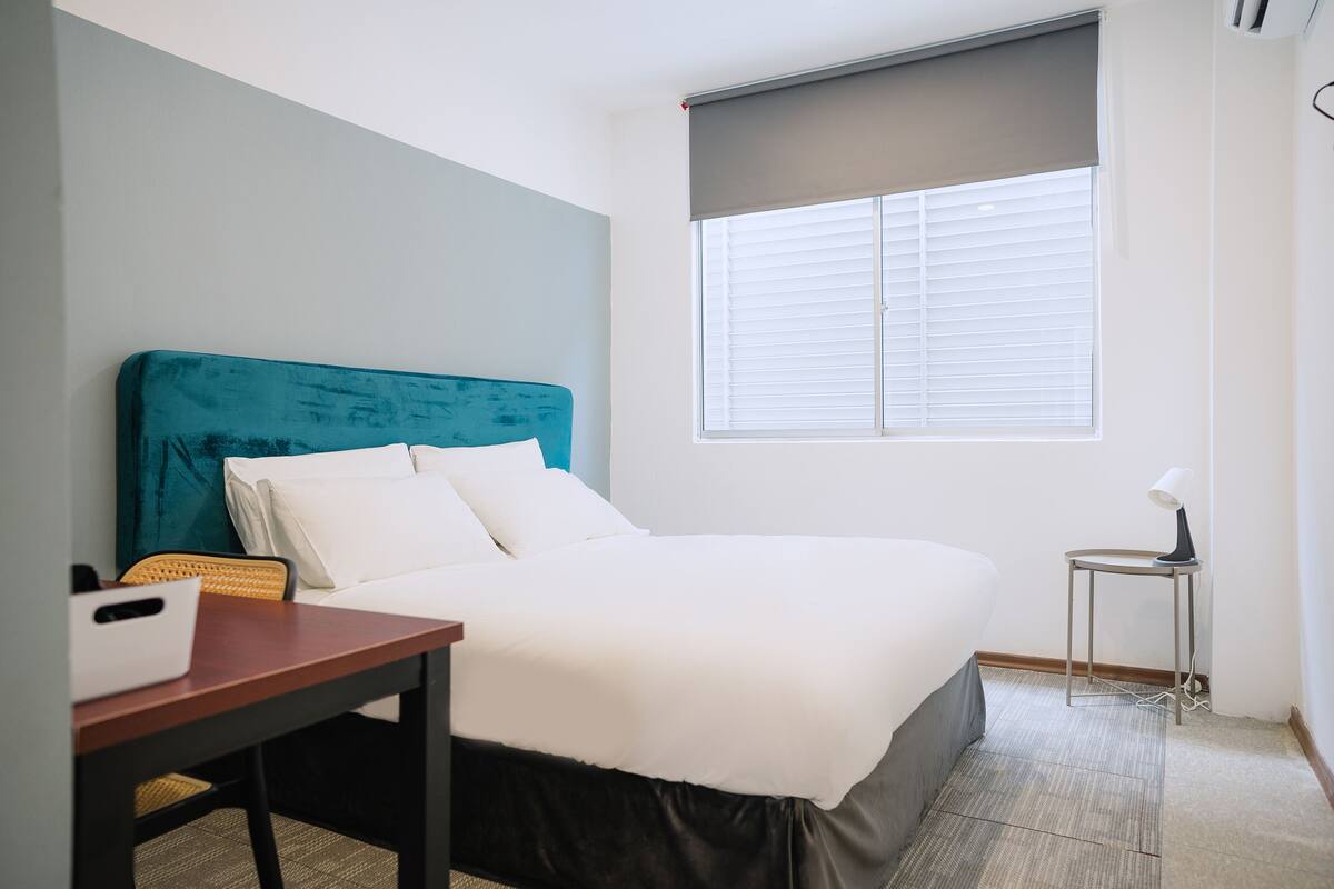 Une chambre d’hôtel bien rangée, avec un lit, une petite table, une chaise et une fenêtre équipée de stores.