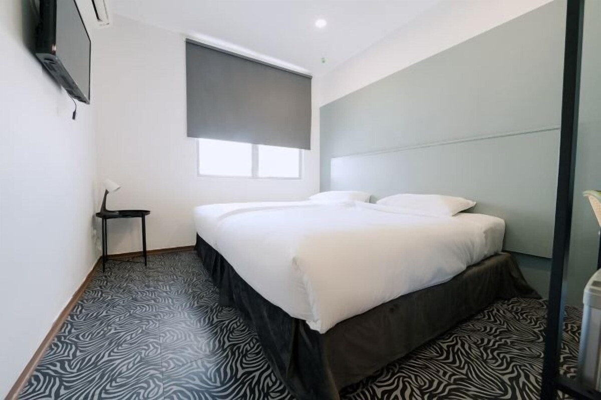 A modern hotel room with a large bed, a small side table, a TV mounted on the wall, and a window with a blind.