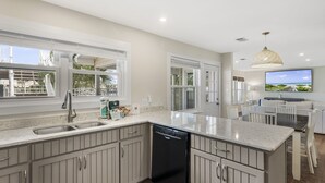 A modern kitchen with a dishwasher, a large island, and a dining area with a TV. - 5BR Waterfront Retreat with Pool, Boat Slips + Dock | Boater’s Dream House (Destin)