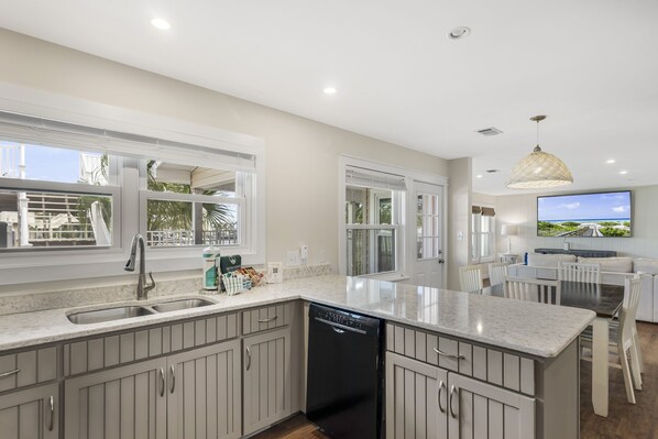 A modern kitchen with a dishwasher, a large island, and a dining area with a TV. - 5BR Waterfront Retreat with Pool, Boat Slips + Dock | Boater’s Dream House (Destin)