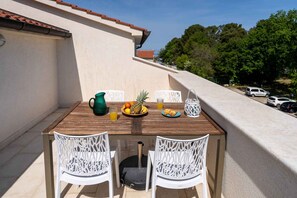 Une table en bois avec des chaises blanches sur un balcon, une carafe, un ananas, des oranges et un verre de jus.