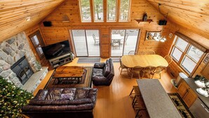 A cozy cabin interior with a stone fireplace, leather sofa, wooden dining table, and large windows offering views of the surrounding forest.