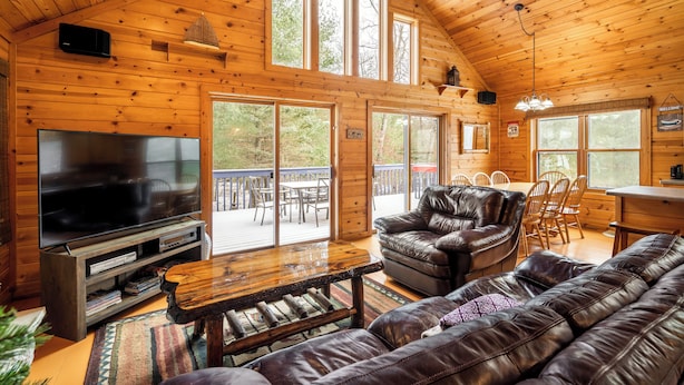 A wooden cabin interior with a large TV, leather sofas, a dining area, and a balcony with a view.